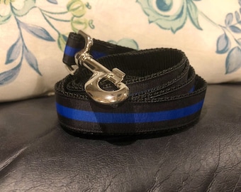 May include: A coiled black nylon dog leash featuring a thin, horizontal royal blue stripe running through the center. The leash is equipped with a polished, silver-toned metal swivel snap hook attached to one end for secure fastening to a collar.