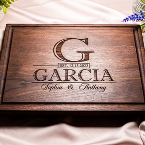 May include: A personalized walnut wood cutting board with the last name "Garcia" engraved in a serif font. The initial "G" is in a larger font and the date "EST. 12.15.2023" is engraved below. The names "Sophia & Anthony" are engraved in a cursive font below the last name.