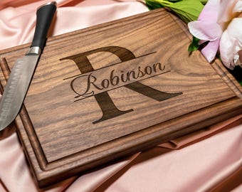 Puede incluir: Una tabla de cortar rectangular de madera marrón oscuro con una letra R grande grabada profundamente y el nombre Robinson escrito en cursiva en el centro. La tabla tiene un borde ranurado y se acompaña de un cuchillo de cocina plateado.