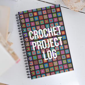 Könnte beinhalten: Ein spiralgebundenes Notizbuch mit einem farbenfrohen Häkelmuster auf dem Einband. Das Cover zeigt ein sich wiederholendes Muster aus bunten Quadraten auf schwarzem Hintergrund. Die Worte "CROCHET PROJECT LOG" sind in Weiß gedruckt, ideal für Häkelprojekte.