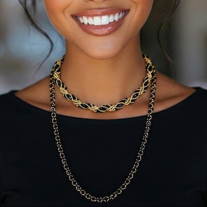 Könnte beinhalten: Zwei Halsketten, eine kurze und eine lange, mit schwarzen und goldenen Perlen. Die kürzere Halskette hat ein gewebtes Design, während die längere ein einfaches Kettenmuster hat. Die Halsketten werden über einem schwarzen Oberteil getragen.