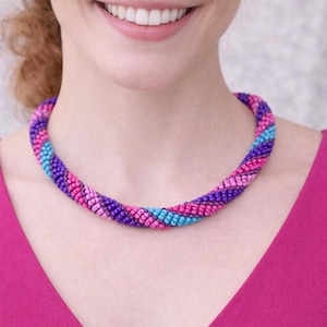 May include: A colorful beaded necklace in shades of pink, purple, and blue. The necklace is a twisted rope design, with a gradient of colors. The necklace is worn around the neck. The background is a light gray.