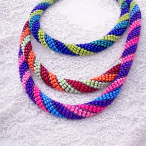 Pode incluir: Três colares de contas coloridas em tons de azul, verde, rosa, roxo, laranja e vermelho. Os colares estão dispostos sobre uma superfície de renda branca, mostrando suas cores vibrantes e o intrincado trabalho de contas.
