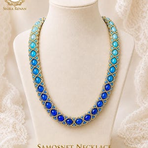 May include: A beaded necklace featuring an ombre gradient of blue cabochon stones, ranging from light turquoise to deep royal blue, set within a gold-toned metallic bead framework. The piece is labelled Samosnet Necklace by Shira Renan.