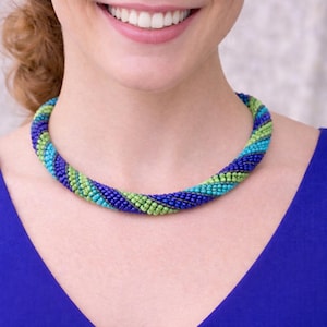 May include: A beaded necklace in shades of blue, green, and turquoise. The necklace is a twisted rope design, with the beads tightly woven together. The necklace is worn against a blue top.
