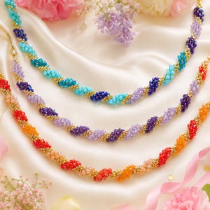 May include: Three beaded necklaces in shades of red, purple, and blue, with gold accents. The necklaces are arranged on a white satin surface, with flowers in the background. The beads are small and closely spaced, creating a textured look.