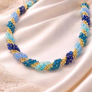 May include: A necklace with a spiral design, made of blue beads and small gold-coloured beads. The beads range in colour from light to dark blue. The necklace is displayed on a white, silky fabric.