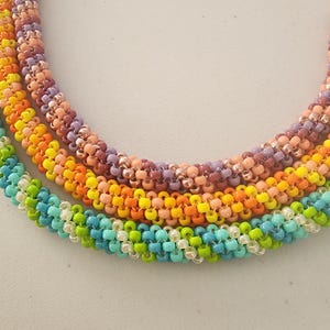 Könnte beinhalten: Ein Set aus fünf bunten Perlenketten, jede mit einem anderen Farbmuster. Die Ketten bestehen aus kleinen, runden Perlen in den Farben Türkis, Grün, Gelb, Orange, Rosa und Lila.