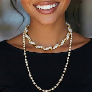 Puede incluir: Collar de dos hilos con cuentas blancas y detalles dorados. El hilo más corto tiene un diseño trenzado, mientras que el hilo más largo es un estilo simple de cuentas. El collar se usa sobre una blusa negra.