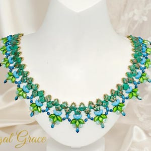 Könnte beinhalten: Eine Halskette mit türkisfarbenen, grünen und blauen Akzenten. Die Halskette wird auf einer weißen Büste ausgestellt. Das Design zeigt ein sich wiederholendes Muster aus türkisfarbenen Steinen, grünen Blättern und blauen Perlen mit goldfarbenen Akzenten.