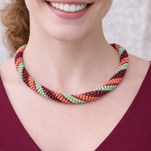 May include: A beaded necklace with a spiral design. The necklace features alternating sections of orange, burgundy, and light green beads. The necklace is worn against a burgundy-colored top.