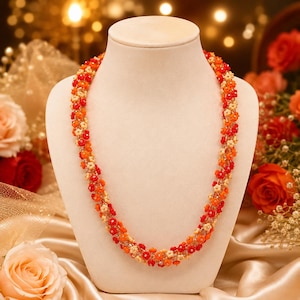 May include: A beaded necklace displayed on a mannequin. The necklace features a floral design with red, orange, and cream-colored beads. The beads are arranged in a spiral pattern. The background includes flowers and a satin fabric.