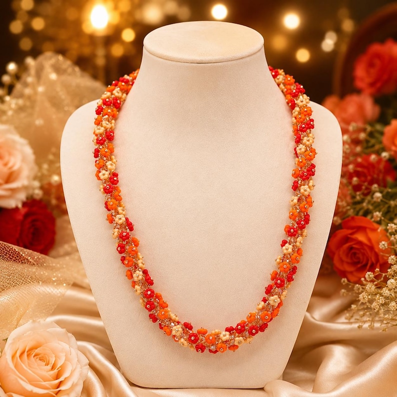 May include: A beaded necklace displayed on a mannequin. The necklace features a floral design with red, orange, and cream-colored beads. The beads are arranged in a spiral pattern. The background includes flowers and a satin fabric.