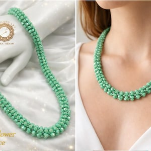 Può includere: Una collana a fiori di cristallo verde menta. La collana è composta da piccole perline sfaccettate, che creano un design floreale strutturato. La collana è esposta su una superficie bianca e indossata attorno al collo di una persona. Il testo "Crystal Flower Necklace" è visibile.