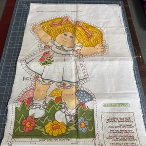 May include: A Cabbage Patch Kids fabric panel featuring a doll with blonde pigtails, wearing a white dress with floral accents. The doll stands in a grassy area with colorful flowers and mushrooms. Instructions are printed on the side.