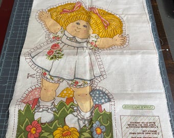 May include: A Cabbage Patch Kids fabric panel depicting a doll with blonde pigtails, dressed in a white dress with floral details. The doll is set in a grassy scene with flowers and mushrooms. Instructions are printed on the side.