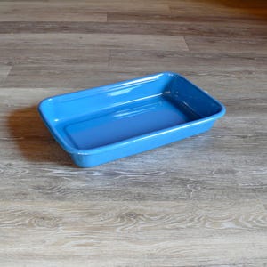 May include: A rectangular, bright blue ceramic baking dish. The dish has slightly rounded corners and a glossy finish. The empty dish is set on a wooden surface.