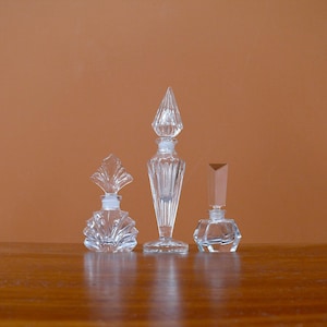 May include: Three clear glass perfume bottles with decorative stoppers. The bottles are arranged in a row on a wooden surface.