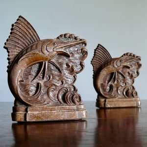 May include: A pair of wooden bookends featuring a carved marlin fish leaping out of the water. The bookends are a dark brown color and have a detailed design.
