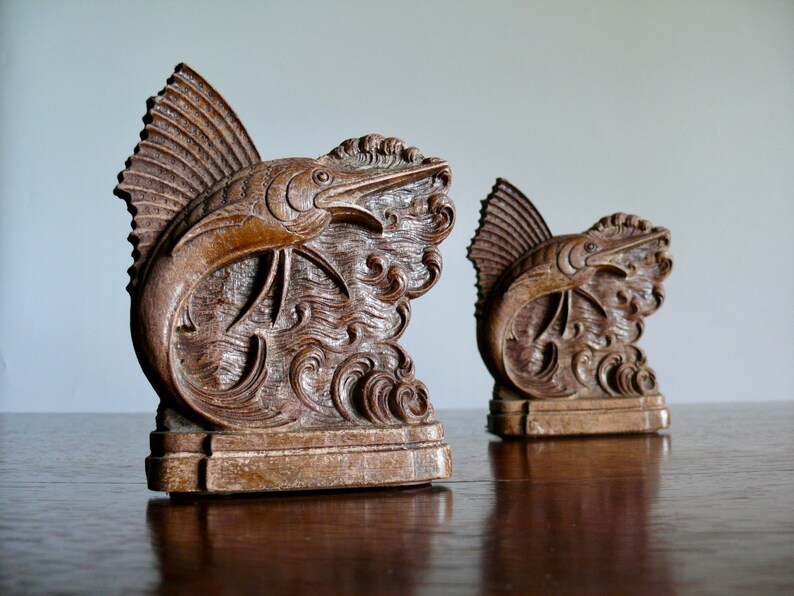 May include: A pair of wooden bookends featuring a carved marlin fish leaping out of the water. The bookends are a dark brown color and have a detailed design.