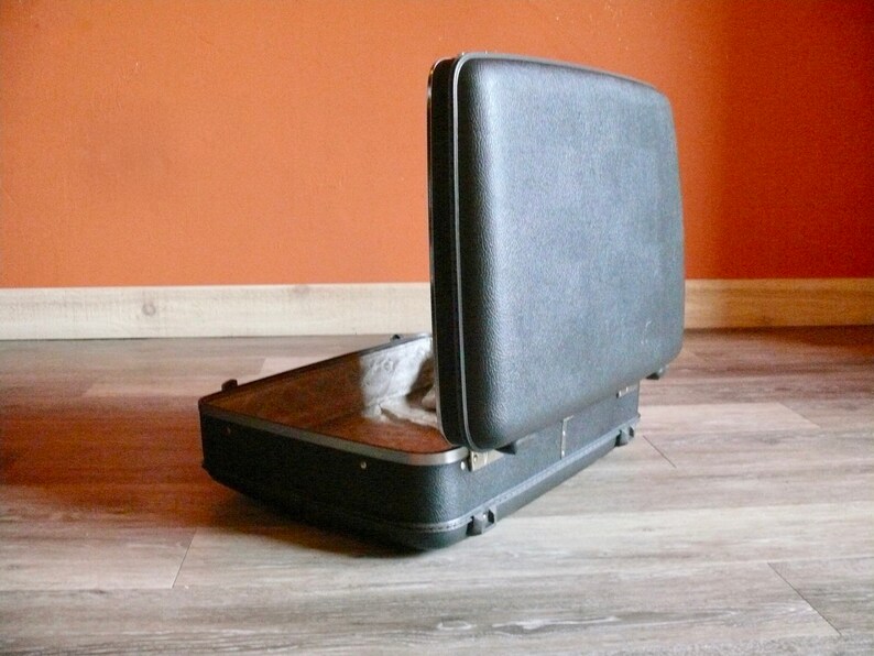 May include: A black hard-shell case with a hinged lid open, revealing an empty interior. The case is sitting on a wood floor.