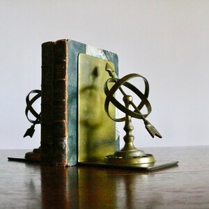 May include: A pair of brass bookends featuring a globe and arrow design. The bookends are holding up a vintage book with a dark green cover.