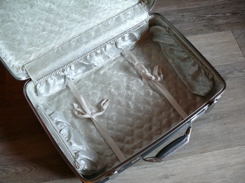 May include: A silver suitcase with a white satin lining and two white satin straps. The suitcase is open and empty.