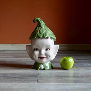 May include: A vintage ceramic cookie jar shaped like a smiling elf face with a green leaf hat and collar. A green apple sits beside the jar. The jar is white with red lips and rosy cheeks.
