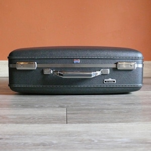 May include: A vintage black hard-sided suitcase with a silver handle and trim. The suitcase has a "Tirma" logo on the front.