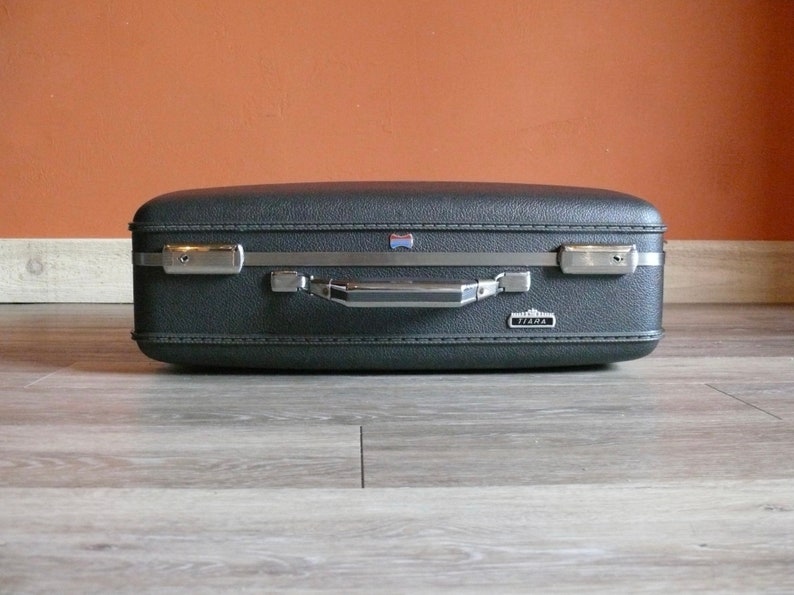 May include: A vintage black hard-sided suitcase with a silver handle and trim. The suitcase has a "Tirma" logo on the front.