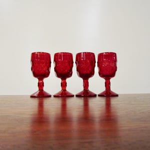 May include: Four small, vibrant red glass goblets arranged in a row. Each goblet features a textured, faceted bowl, a short stem, and a wide, circular base. The glassware is translucent, allowing light to pass through the deep red material.