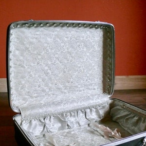 May include: A vintage silver suitcase with a white floral lining. The suitcase is open and the lining is visible.