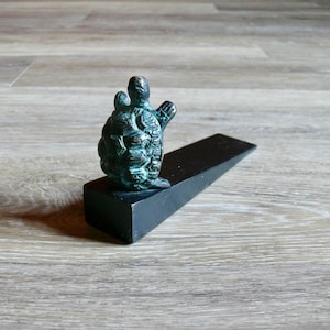 May include: A bronze-colored turtle figurine sits atop a black doorstop. The turtle has a textured shell and is positioned on a wedge-shaped doorstop. The doorstop is made of a dark, solid material.