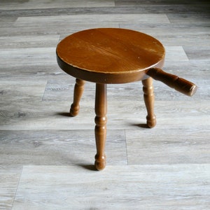 May include: A wooden stool with a round seat and three legs. The stool has a single handle extending from the side of the seat.