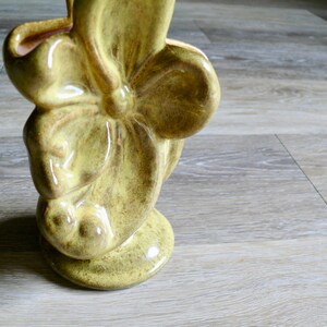 K&ouml;nnte beinhalten: Eine Keramikvase mit floralem Design. Die Vase hat eine ged&auml;mpfte gelbgr&uuml;ne Farbe mit gl&auml;nzendem Finish. Das Design zeigt eine gro&szlig;e, stilisierte Blume mit abgerundeten Bl&uuml;tenbl&auml;ttern und einer strukturierten Oberfl&auml;che. Die Vase hat einen Sockel.