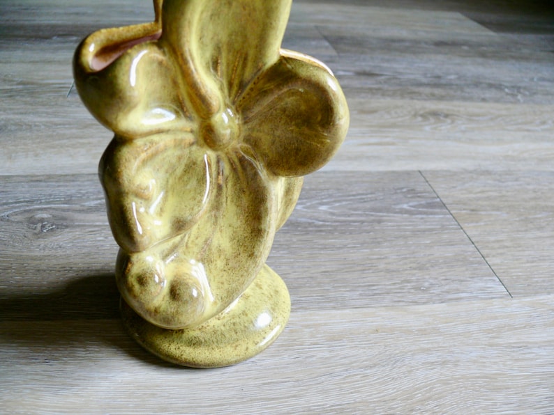 K&ouml;nnte beinhalten: Eine Keramikvase mit floralem Design. Die Vase hat eine ged&auml;mpfte gelbgr&uuml;ne Farbe mit gl&auml;nzendem Finish. Das Design zeigt eine gro&szlig;e, stilisierte Blume mit abgerundeten Bl&uuml;tenbl&auml;ttern und einer strukturierten Oberfl&auml;che. Die Vase hat einen Sockel.