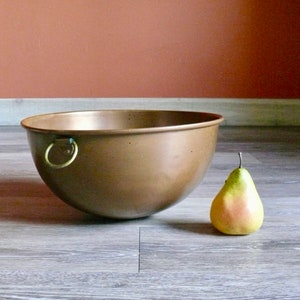May include: A large copper bowl with a ring handle. The bowl is sitting on a wood floor next to a green pear.