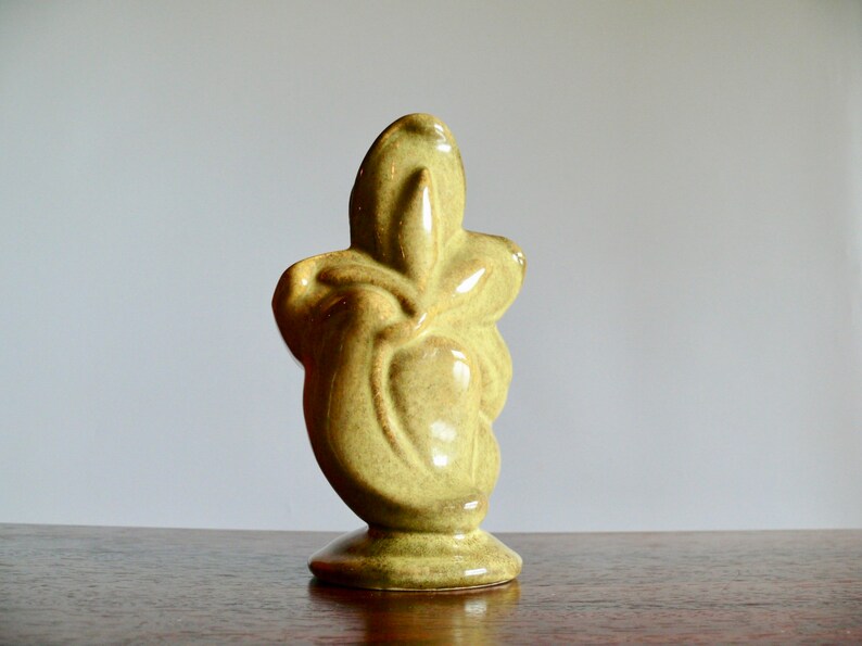 K&ouml;nnte beinhalten: Eine hellgr&uuml;ne Keramikskulptur mit Sockel. Die abstrakte Form hat eine glatte, gl&auml;nzende Oberfl&auml;che und eine einzigartige, organische Form. Die Skulptur wird auf einer Holzoberfl&auml;che vor einem neutralen Hintergrund ausgestellt.