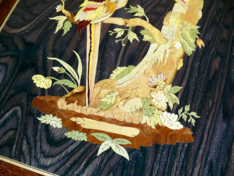 K&ouml;nnte beinhalten: Intricate Holzmarqueterie-Kunstwerk mit einem gelben und roten Vogel, der auf einem Ast mit gr&uuml;nen Bl&auml;ttern und Blumen sitzt. Der Hintergrund ist eine dunkle Holzmaserung.