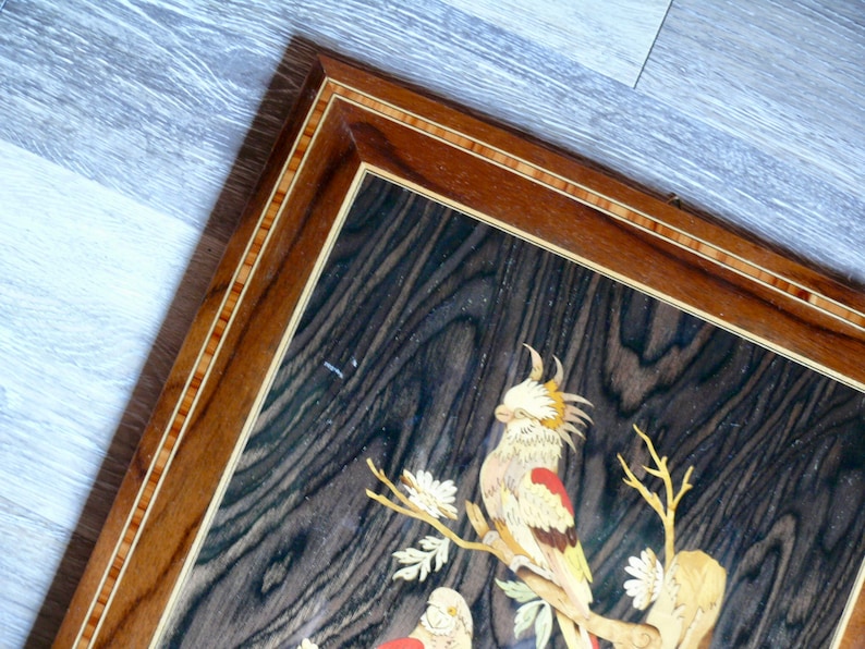 K&ouml;nnte beinhalten: Ein gerahmtes Holz-Marqueterie-Kunstwerk mit einer detaillierten Darstellung von zwei V&ouml;geln, die auf einem Ast mit wei&szlig;en Bl&uuml;ten sitzen. Die V&ouml;gel sind aus verschiedenen Holzst&uuml;cken in verschiedenen Farben gefertigt, was einen Mosaik-Effekt erzeugt. Der Rahmen ist aus dunklem Holz mit einer Einlage aus hellem Holz.