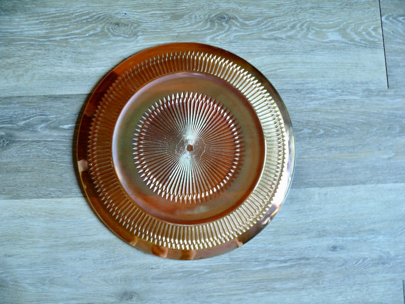 Puede incluir: Un plato de servicio dorado con un dise&ntilde;o decorativo en forma de sol. El plato tiene un borde elevado y una superficie lisa y brillante.