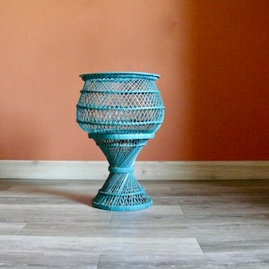 May include: A teal blue wicker plant stand with a wide, round top and a tall, slender base. The stand is made of woven wicker and has a delicate, airy design.
