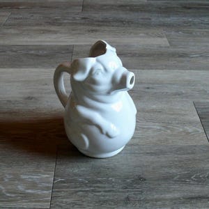 May include: A white ceramic pig-shaped pitcher with a handle and a spout. The pitcher has a detailed design, including a pig's snout, ears, and a curled tail. The pitcher is sitting on a wooden surface.