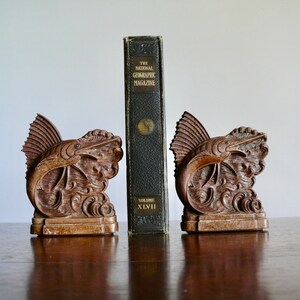 May include: A pair of wooden bookends shaped like a marlin fish leaping out of the water. The bookends are a dark brown color and have intricate details. The bookends are holding up a vintage copy of "The National Geographic Magazine" volume XLVI.