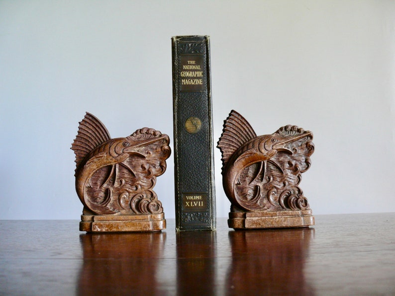 May include: A pair of wooden bookends shaped like a marlin fish leaping out of the water. The bookends are a dark brown color and have intricate details. The bookends are holding up a vintage copy of "The National Geographic Magazine" volume XLVI.