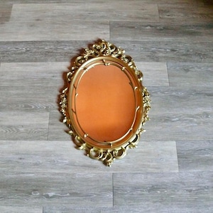 May include: An oval-shaped mirror with a gold-colored ornate frame. The frame has a floral design and is made of plastic.
