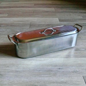 May include: A stainless steel rectangular roasting pan with a lid. The lid has a handle and a brown interior. The pan has two handles on the sides. The pan is sitting on a wooden floor.