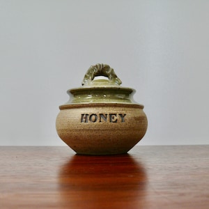 May include: A green and brown ceramic honey pot with a unique handle and the word "Honey" inscribed on the front.