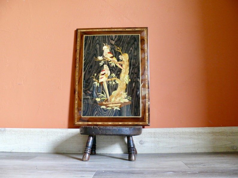 K&ouml;nnte beinhalten: Ein gerahmtes Holz-Marqueterie-Kunstwerk, das drei wei&szlig;e V&ouml;gel mit roten Akzenten zeigt, die auf einem Ast sitzen. Das Kunstwerk wird auf einem Holzhocker ausgestellt.