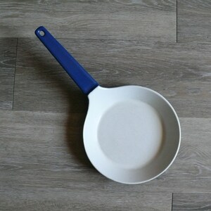 May include: A white ceramic frying pan with a blue handle. The pan is empty and sitting on a wood surface.